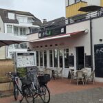Al Parco Norderney: Italienisch essen mit Terrasse und Strandkorb Al Parco Norderney – Fassade mit Schild „Café Ristorante Pizzeria“, Veltins-Ausschank, Außenterrasse mit Strandkorb und Tafeln „Warme Küche ab 11:00“ sowie „Fleisch, Fisch, Nudeln, Salate, Pizza, Suppen“