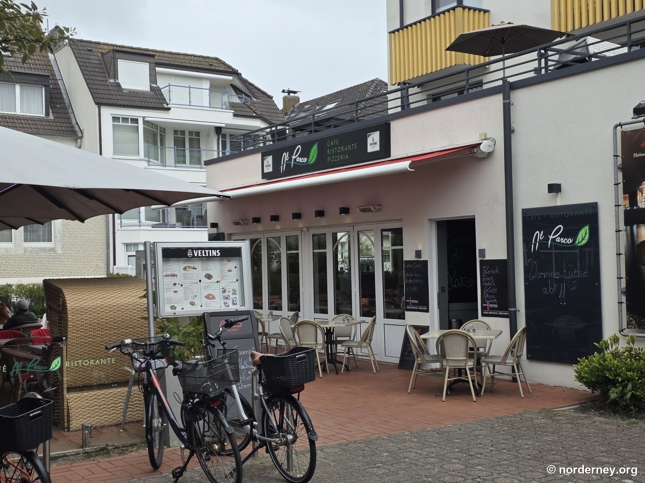 Al Parco Norderney – Fassade mit Schild „Café Ristorante Pizzeria“, Veltins-Ausschank, Außenterrasse mit Strandkorb und Tafeln „Warme Küche ab 11:00“ sowie „Fleisch, Fisch, Nudeln, Salate, Pizza, Suppen“