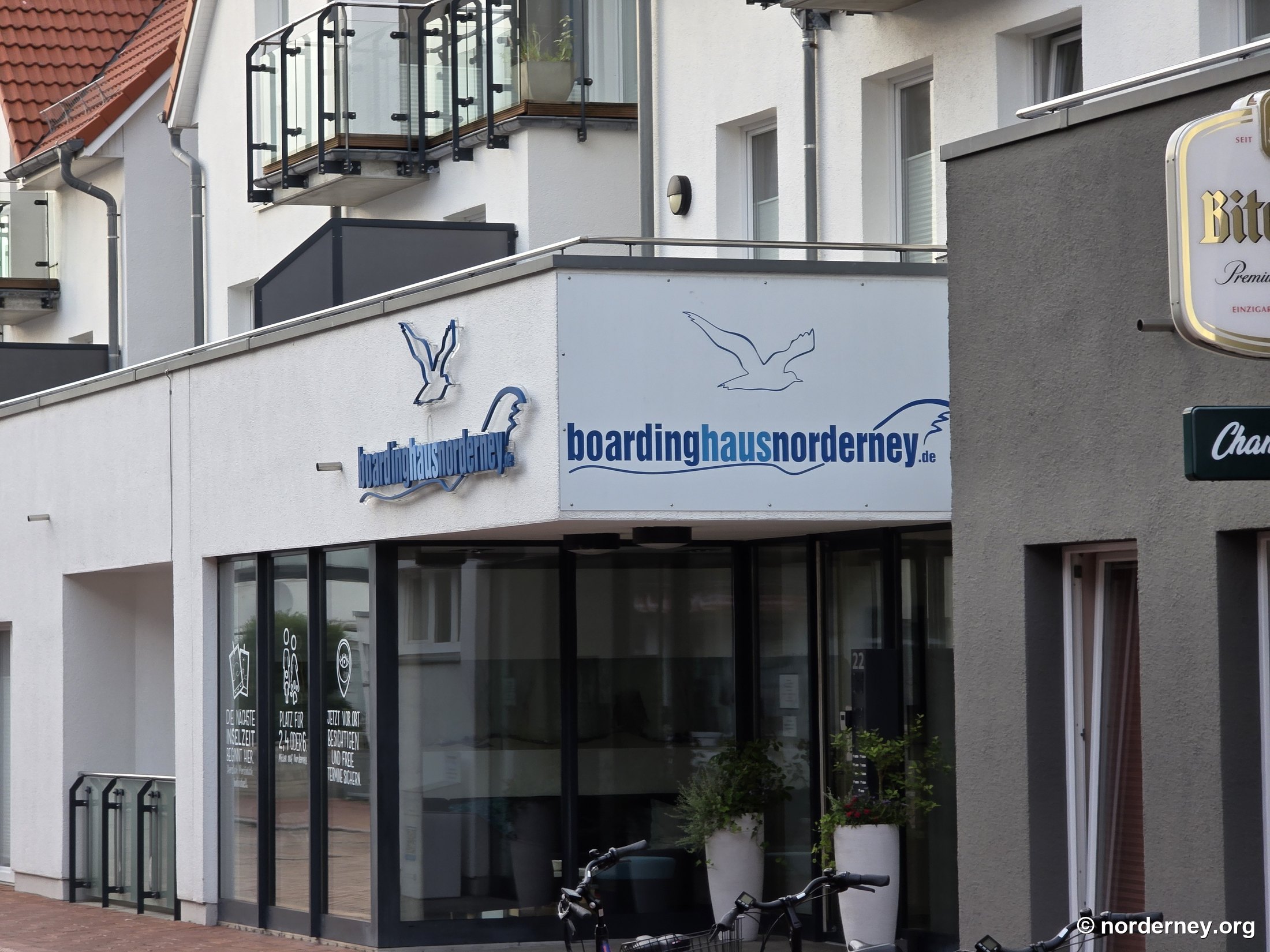 Helle Eckfassade des Boardinghauses mit Möwen-Logo, Schriftzug „boardinghausnorderney.de“ und Eingang mit Glasfront Helle Eckfassade des Boardinghauses mit Möwen-Logo, Schriftzug „boardinghausnorderney.de“ und Eingang mit Glasfront