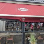 Columbus Norderney: Moderne Inselküche, Bar und Terrasse im Ortskern Columbus Norderney – rote Markise mit Anker-Logo und Terrassenverglasung „Treffen · Erleben · Genießen“ in der Mittelstraße
