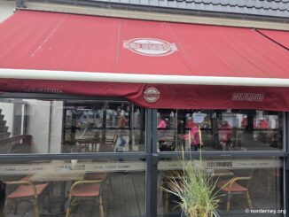 Columbus Norderney – rote Markise mit Anker-Logo und Terrassenverglasung „Treffen · Erleben · Genießen“ in der Mittelstraße