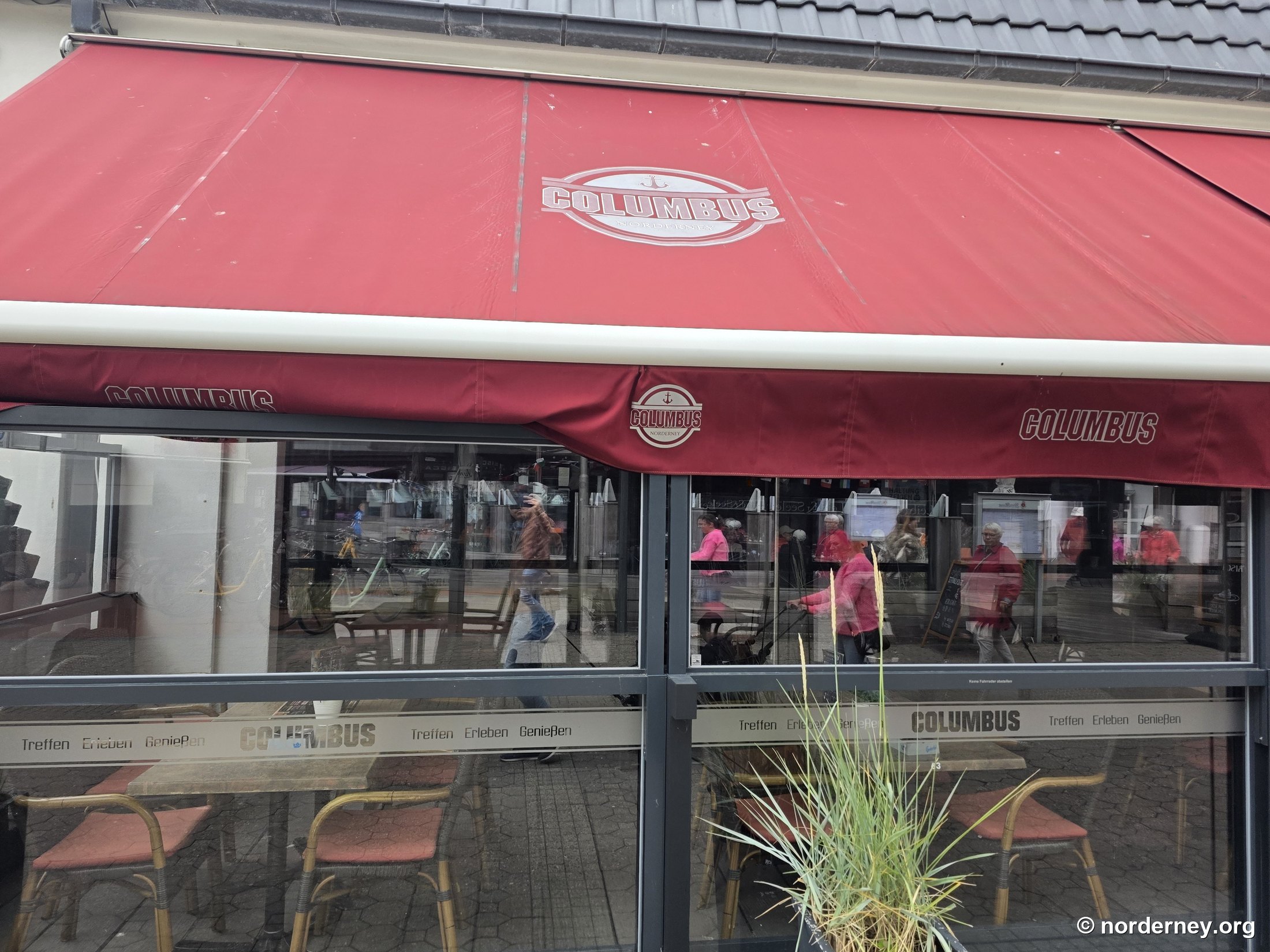 Columbus Norderney – rote Markise mit Anker-Logo und Terrassenverglasung „Treffen · Erleben · Genießen“ in der Mittelstraße