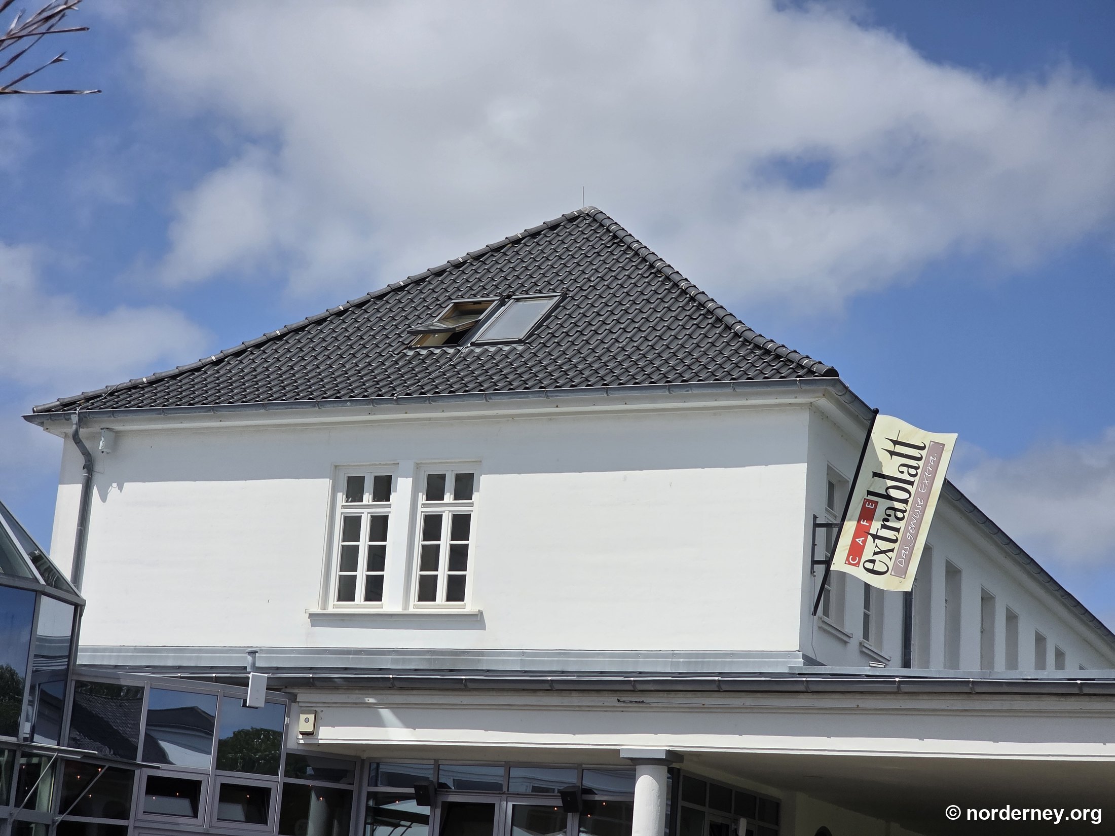 Café Extrablatt Norderney – Giebel mit Ausleger-Fahne „CAFE Extrablatt – Das Genuss Extra“ und verglastem Wintergarten-Anbau am Kurplatz
