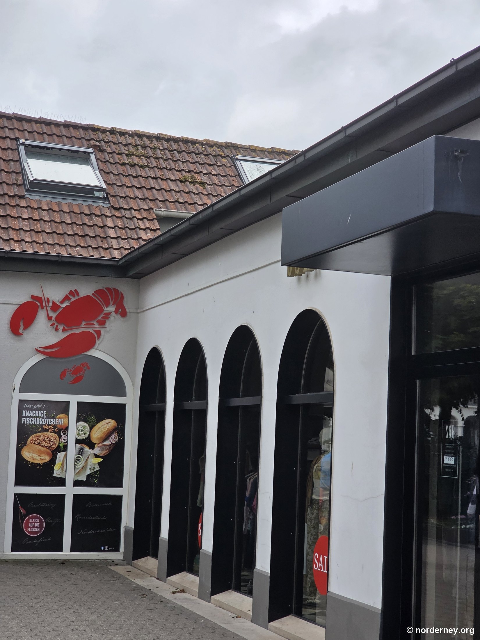 Gosch Norderney – Fassade am Kurplatz mit rotem Hummer-Logo und Plakat „Hier gibt's knackige Fischbrötchen!“
