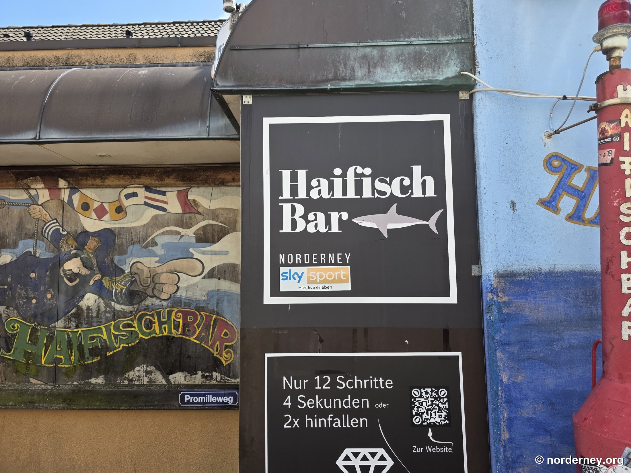 Werbeschild der Haifischbar Norderney mit Sky-Sport-Hinweis neben dem Straßenschild „Promilleweg“