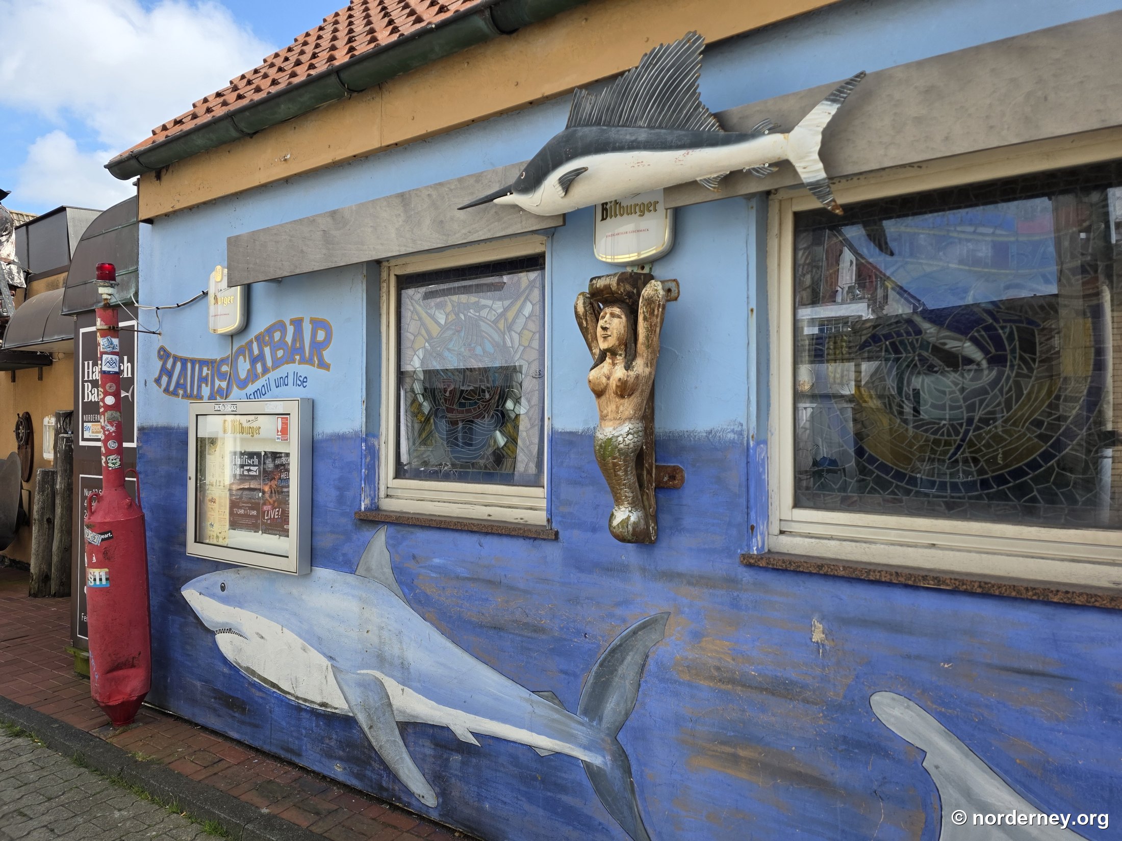 Blaue Fassade der Haifischbar Norderney mit großem Hai-Wandbild, hölzernem Schwertfisch, Galionsfigur, Schaukasten und rotem Poller