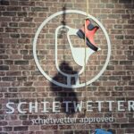 Schietwetter Norderney: Mode und Regenkleidung an vier Standorten Schietwetter „schietwetter approved“ – Möwen-Logo mit rotem Gummistiefel an einer Backsteinwand im Laden auf Norderney