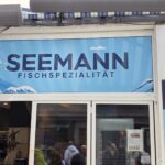 Seemann Norderney: Fischspezialitäten in der Poststraße Seemann Fischspezialität in der Poststraße auf Norderney – Ladenfront mit Wellen-Logo, Anker-Emblem „Since 2025“ und Speisekarte im Schaufenster