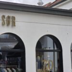 SØR Norderney: Gehobene Damen- und Herrenmode am Kurplatz SØR Norderney – Fassade am Kurplatz mit goldenem Schriftzug, drei Rundbogenfenstern und Jubiläums-Dekoration „70 Jahre“ im Schaufenster