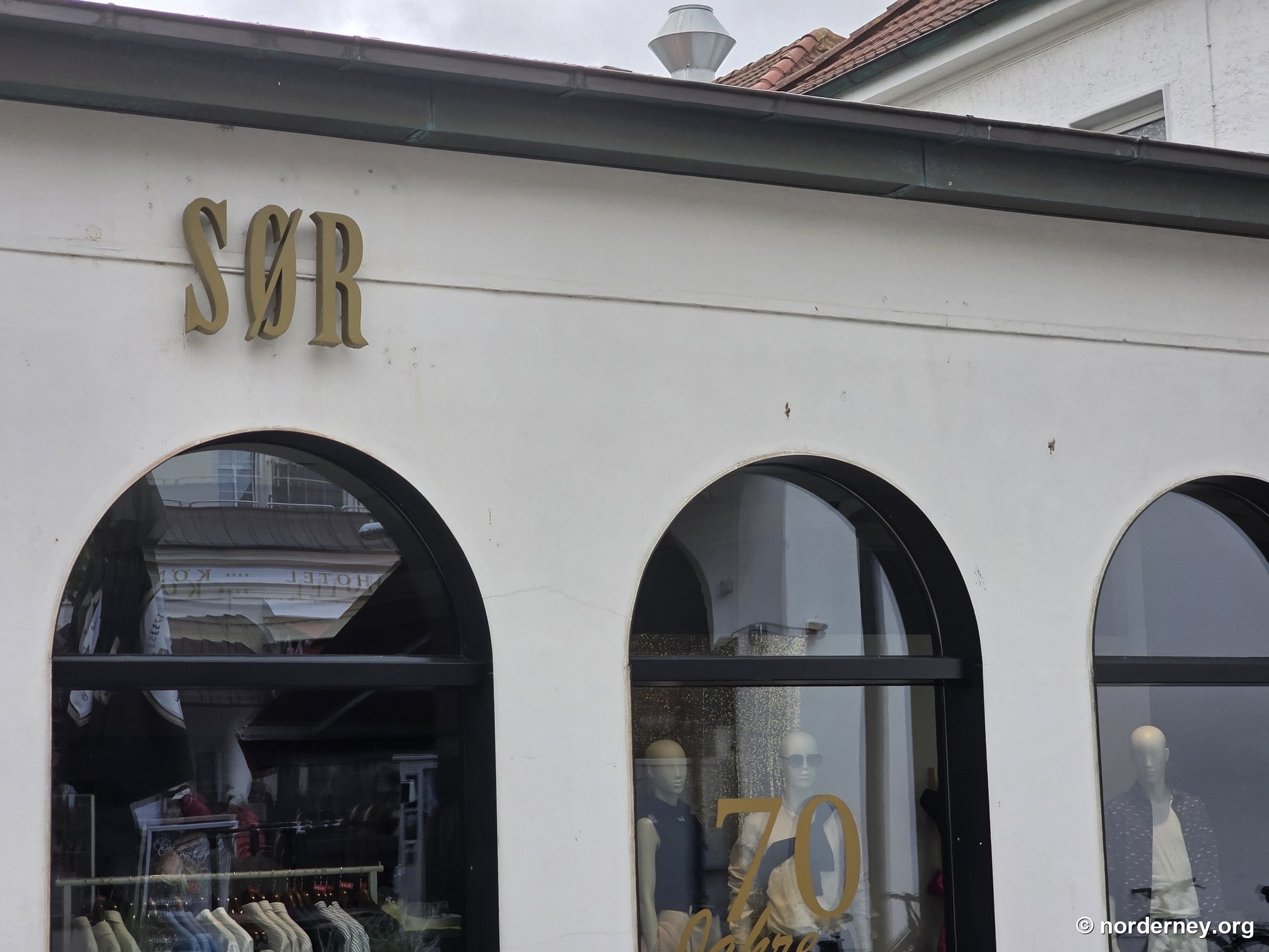 SØR Norderney – Fassade am Kurplatz mit goldenem Schriftzug, drei Rundbogenfenstern und Jubiläums-Dekoration „70 Jahre“ im Schaufenster