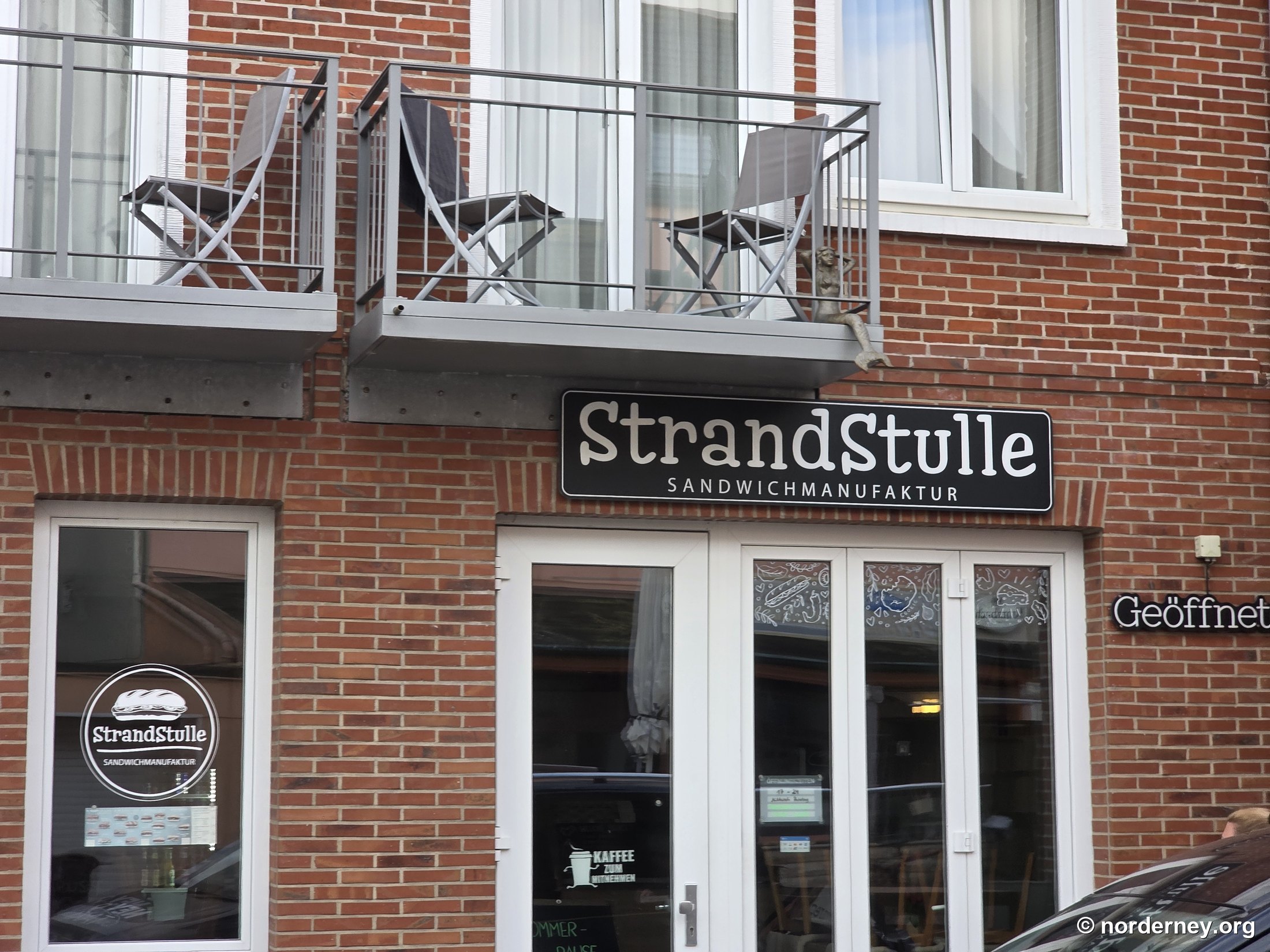 Backstein-Fassade der StrandStulle in der Jann-Berghaus-Straße mit Sandwichmanufaktur-Schild, Balkon und Leuchtschild „Geöffnet“ Backstein-Fassade der StrandStulle in der Jann-Berghaus-Straße mit Sandwichmanufaktur-Schild, Balkon und Leuchtschild „Geöffnet“