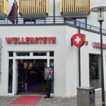 Wellensteyn Norderney: Jacken-Store am Kurplatz Wellensteyn-Store auf Norderney – Fassade in der Adolfsreihe mit rotem Schriftzug-Logo, weiß-rotem Wappen und rotem Teppich am Eingang