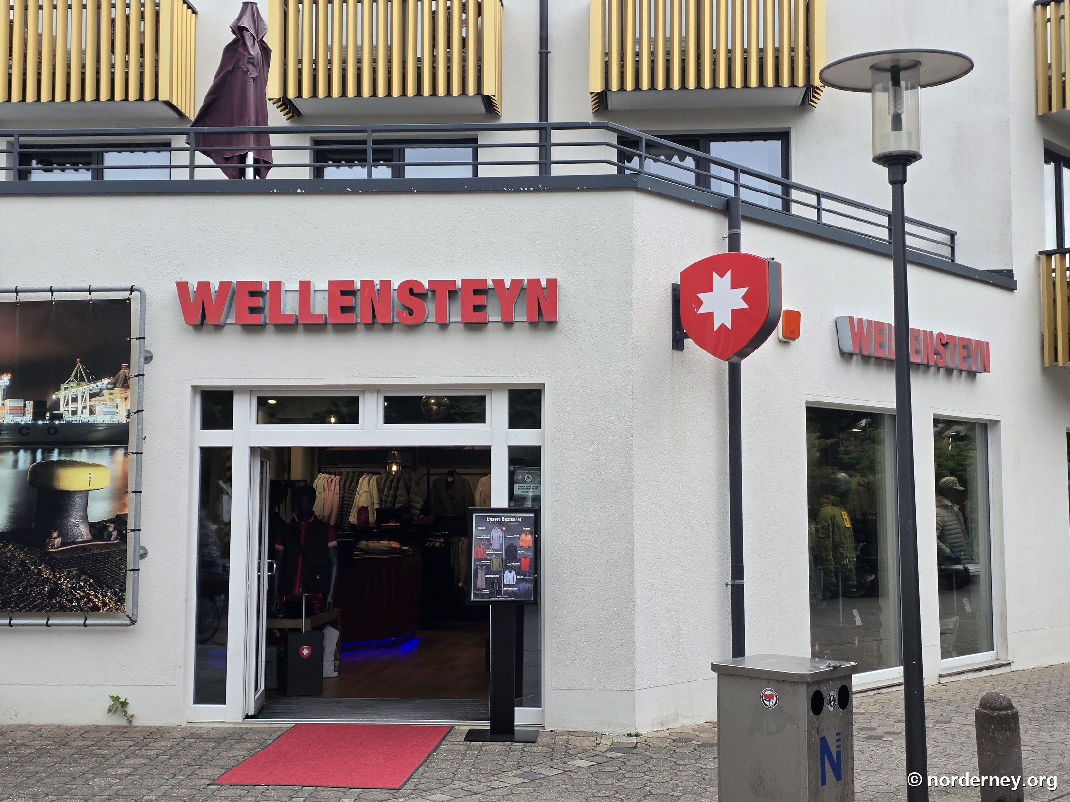 Wellensteyn-Store auf Norderney – Fassade in der Adolfsreihe mit rotem Schriftzug-Logo, weiß-rotem Wappen und rotem Teppich am Eingang