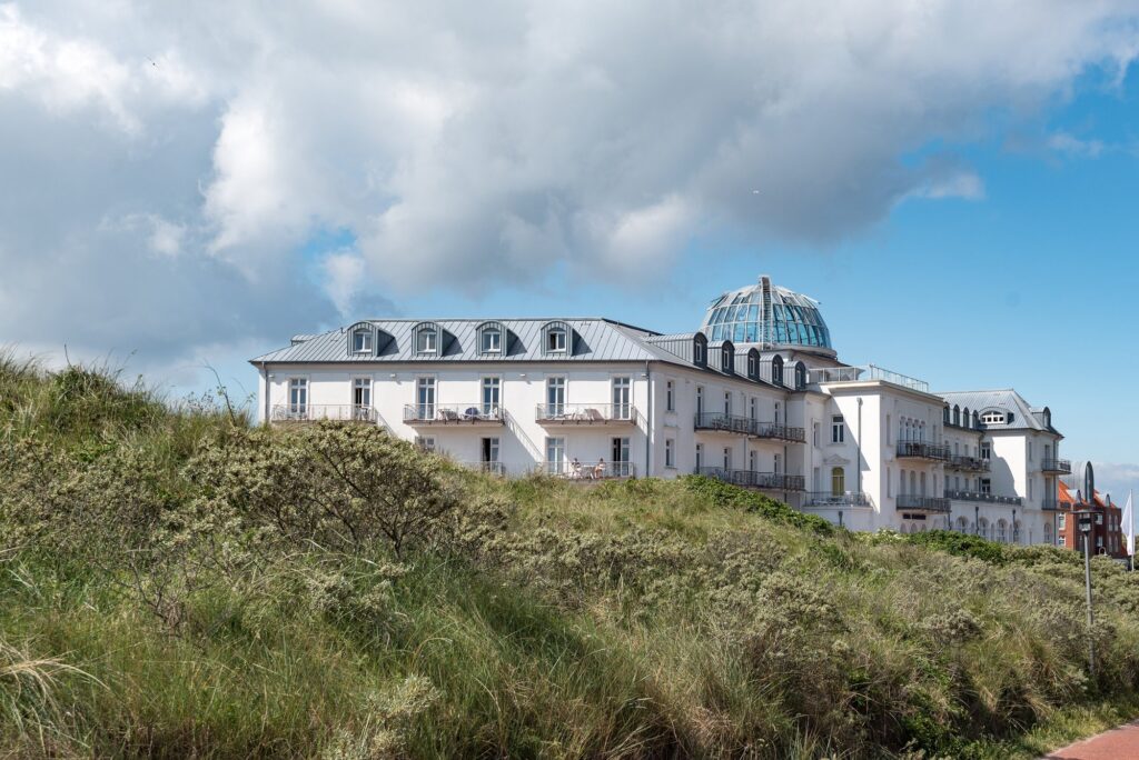 Weißes Strandhotel Kurhaus Juist mit Glaskuppel hinter bewachsenen Dünen an der Strandpromenade