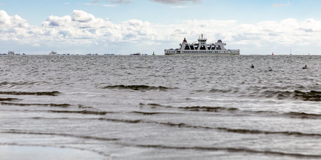 Frisia-Fähre auf dem Wattenmeer vor Norderney unter Wolken, im Vordergrund Wellen