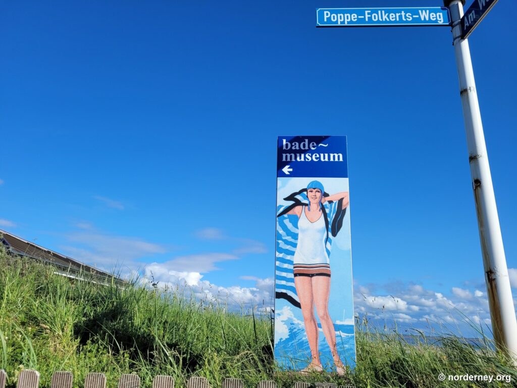 Wegweiser zum Bademuseum Norderney mit Straßenschild Poppe-Folkerts-Weg vor Dünengras